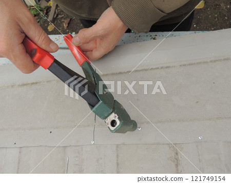 top view of carpenter's hands holding rivet gun and fixing staples into sheet metal, fastening iron flat parts with riveter top angle view 121749154