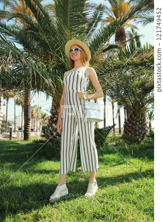 Summer outfit, stylish beautiful young woman posing in white striped jumpsuit in park, palm trees Summer outfit, stylish beautiful young woman posing in white striped jumpsuit in park, palm trees 121749452