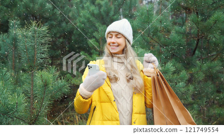 Happy smiling woman with phone, shopping bag in winter against Christmas tree, girl using smartphone 121749554