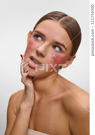 Portrait of young female model in dreamy pose with sparkling golden accents on pink cheeks against white studio background. Individuality. Concept of modern beauty. Portrait of young female model in dreamy pose with sparkling golden accents on pink cheeks against white studio background. Individuality. Concept of modern beauty. 121750300