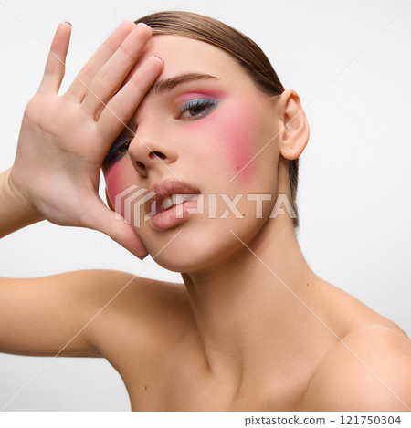 Close-up of model with vibrant pink blush and pastel eye makeup, gracefully covering part of her face with hand against white studio background. Concept of natural beauty. Close-up of model with vibrant pink blush and pastel eye makeup, gracefully covering part of her face with hand against white studio background. Concept of natural beauty. 121750304