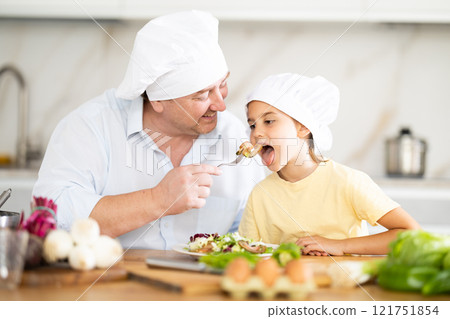 Father in a chef hat offers his little daughter to try the prepared salad in kitchen Father in a chef hat offers his little daughter to try the prepared salad in kitchen 121751854