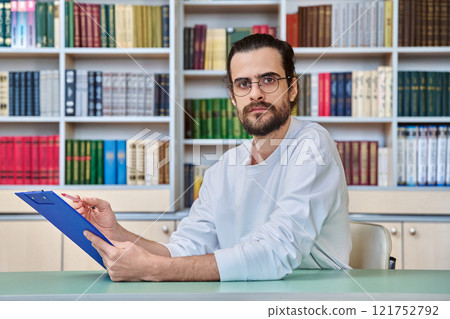 Portrait of young male teacher, psychologist with clipboard sitting at desk in library Portrait of young male teacher, psychologist with clipboard sitting at desk in library 121752792
