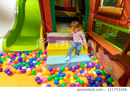 A cheerful indoor playground with a child playing near a colorful ball pit and slide. 121753268