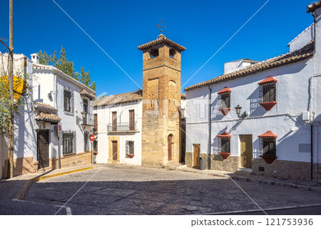 Ronda town in Spain. Historic town square with a distinctive stone tower and whitewashed buildings under a vibrant blue sky. 121753936