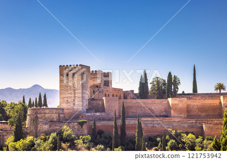 View of the Alhambra palace from the Mirador de San Nicolas lookout in Granada town in Spain. Ancient stone fortress with tall trees against a clear blue sky. 121753942