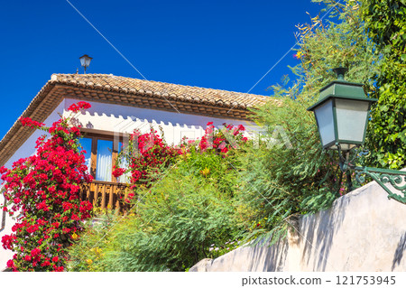 Granada town in Spain. Vibrant flowers cascade down a white building's facade, creating a picturesque scene against a clear blue sky. Granada town in Spain. Vibrant flowers cascade down a white building's facade, creating a picturesque scene against a clear blue sky. 121753945