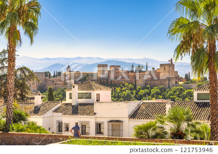 The Alhambra palace from viewpoint in Granada town in Spain. Historic castle viewed from a sun-drenched hillside, palm trees framing the scene. A person observes the view. 121753946