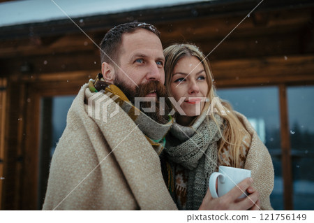Couple bundled in thick blanket standing outdoor, hugging in front of a cabin during snowfall. Couple bundled in thick blanket standing outdoor, hugging in front of a cabin during snowfall. 121756149