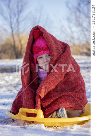A smiling child wrapped in a cozy red blanket, sitting on a yellow sled in a snowy outdoor setting, enjoying a crisp winter day. A smiling child wrapped in a cozy red blanket, sitting on a yellow sled in a snowy outdoor setting, enjoying a crisp winter day. 121758124