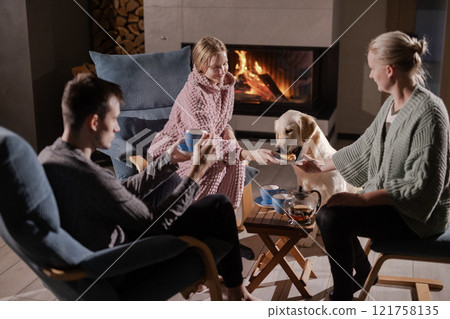 Group of friends relaxing by a fireplace, enjoying tea and snacks, with a cheerful Golden Retriever in a warm and inviting home setting. 121758135