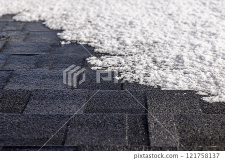 Close-up of a snow-dusted asphalt shingle roof with sunlight highlighting the texture and geometric pattern of the tiles. 121758137