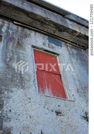 A stone building with a red door on an old deserted property. Architecture details of the exterior of a grey industrial built structure. Weathered concrete walls with a high up wooden doorway 121759480