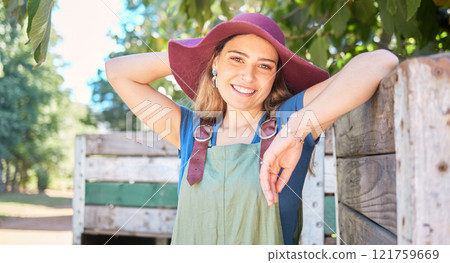 Portrait of a female farmer standing under a tree next to a rustic wooden crate on a organic and sustainable farm. One young happy woman smiling outside in an orchard on a sunny day picking apples Portrait of a female farmer standing under a tree next to a rustic wooden crate on a organic and sustainable farm. One young happy woman smiling outside in an orchard on a sunny day picking apples 121759669