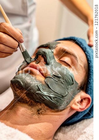 A man relaxes as a professional applies a green clay face mask, showing the tranquil environment of a spa during an afternoon treatment A man relaxes as a professional applies a green clay face mask, showing the tranquil environment of a spa during an afternoon treatment 121762018