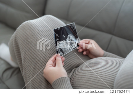Gen Z pregnant woman holding ultrasound scan photo of a fetus sitting on a sofa. Concept of pregnancy, Maternity prenatal care Gen Z pregnant woman holding ultrasound scan photo of a fetus sitting on a sofa. Concept of pregnancy, Maternity prenatal care 121762474