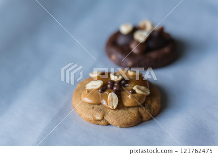 Close up of homemade american traditional chocolate chip cookie with peanut butter, caramel and peanuts topping. Dark chocolate cookie on the background. Close up of homemade american traditional chocolate chip cookie with peanut butter, caramel and peanuts topping. Dark chocolate cookie on the background. 121762475