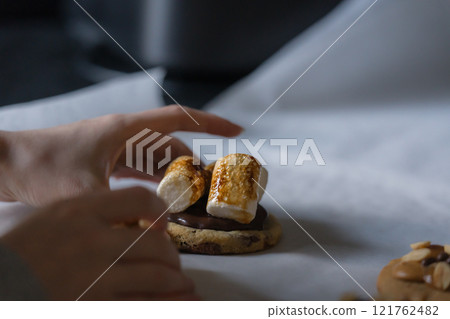 Woman hand taking homemade american chocolate chip cookie with burnt marshmallows from white baking paper. Woman hand taking homemade american chocolate chip cookie with burnt marshmallows from white baking paper. 121762482