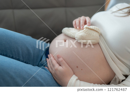 Pregnant woman holding small baby shoes on her belly relaxing at home on couch 121762487