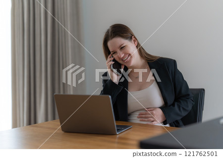 Cheerful gen z pregnant woman working on laptop at home and talking to the phone.  121762501