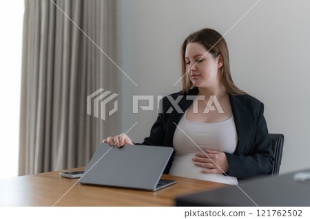Gen Z pregnant business woman using a laptop and holding one hand on her belly while working at home office remotely Gen Z pregnant business woman using a laptop and holding one hand on her belly while working at home office remotely 121762502