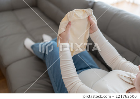Pregnant woman holding baby onesie bodysuit sitting on a sofa. Preparing and planning baby clothes for new baby, packing for maternity at home  121762506