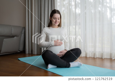 Caucasian young pregnant woman holding her belly while sitting on yoga mat at home. Workout, yoga and fitness at home, pregnancy concept. Pre-natal. 121762519