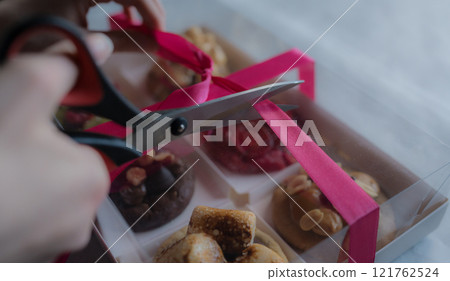 Woman opens gift box of festive decorated homemade cookies with toppings using scissors. Berries, pistachio, caramel, marshmallow, peanuts, chocolate Woman opens gift box of festive decorated homemade cookies with toppings using scissors. Berries, pistachio, caramel, marshmallow, peanuts, chocolate 121762524