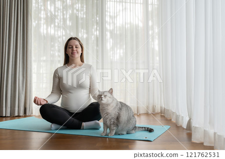 Young pregnant woman with cute Scottish Straight cat meditating on yoga mat at home, eyes closed. Concept of yoga during pregnancy. High quality photo 121762531