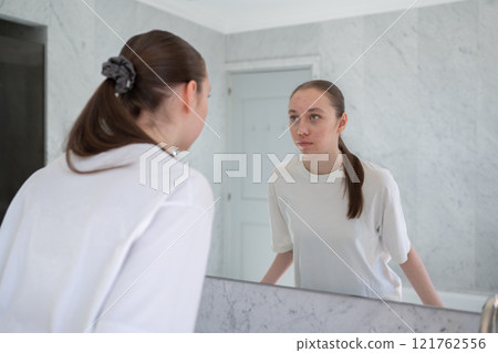 Gen z woman looking at her acne, stands before a mirror in bathroom. Concept of acne treatment, skincare, adult acne 121762556