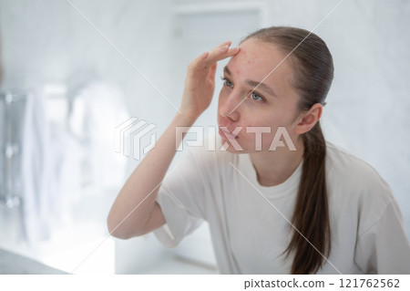 Gen z woman looking at her acne on forehead, stands before a mirror in bathroom touching her skin. Concept of acne treatment, skincare, adult acne 121762562