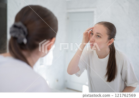 Unhappy gen z woman finding an acne on forehead, stands before a mirror in the bathroom. Concept of acne treatment, skincare, adult acne 121762569