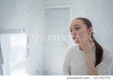 Gen z woman looking at her acne on the cheek, stands before a mirror in bathroom touching her skin. Concept of acne treatment, skincare, adult acne 121762570