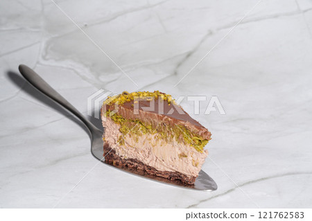 Slice of chocolate Dubai cheesecake with pistachio paste and kataifi covered in chocolate ganache on cake spatula. White background with copy space. Indulgence, gourmet desserts, culinary presentation Slice of chocolate Dubai cheesecake with pistachio paste and kataifi covered in chocolate ganache on cake spatula. White background with copy space. Indulgence, gourmet desserts, culinary presentation 121762583