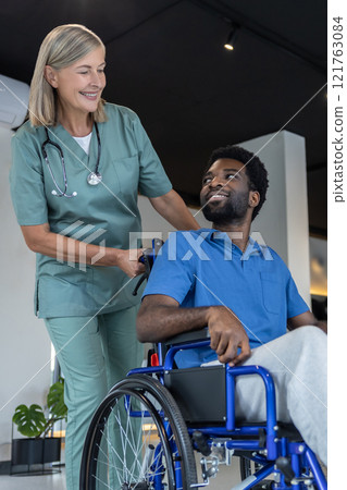 Mature female doctor carrying a wheeling-chair with a male patient 121763084