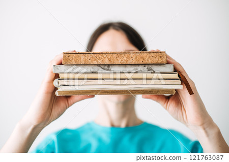 Young woman holding a stack of notebooks with unique textures and materials in a bright minimalist background. Creative journaling and mindful self-expression. 121763087