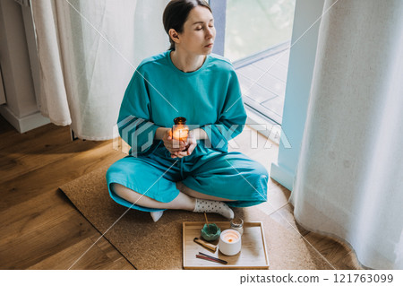 Woman holding candle during a moment of stillness in serene home setting. The ritual of stillness, modern spirituality, and energy cleansing. 121763099