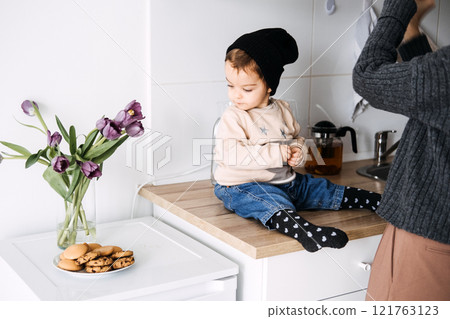Child sitting on kitchen counter with vase of tulips and cookies. Cozy home moment, family kitchen, childhood innocence, domestic life, simple pleasures. 121763123