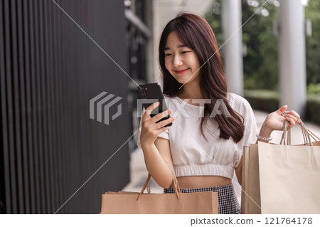 Young Woman Shopping with Paper Bag and Smartphone in Urban Setting, Smiling and Enjoying a Modern Retail Experience Young Woman Shopping with Paper Bag and Smartphone in Urban Setting, Smiling and Enjoying a Modern Retail Experience 121764178