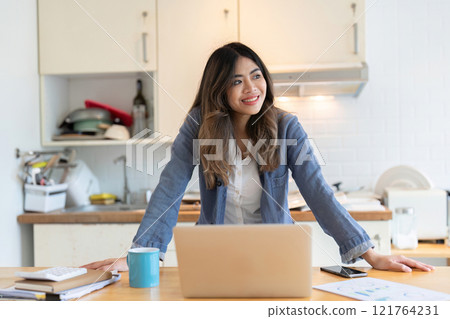 Young Freelance Woman Working from Home in Modern Dining Room with Laptop and Coffee Mug, Smiling and Looking Inspired 121764231