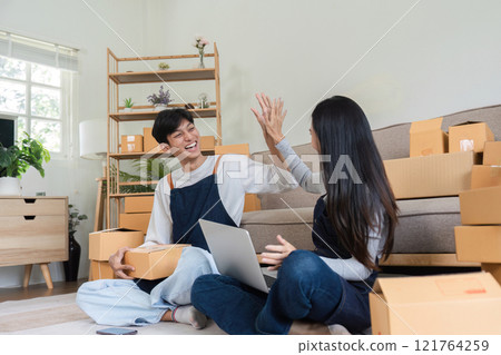 Happy Couple Celebrating Successful Online Sales with High Five in Modern Living Room Surrounded by Shipping Boxes and Laptop 121764259