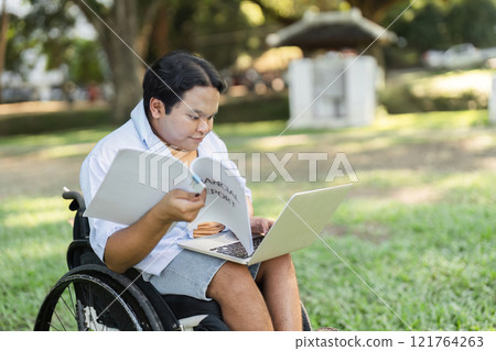Young Disabled Businessman in Wheelchair Analyzing Outdoor Work Environment with Laptop and Documents 121764263