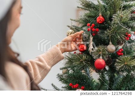 Young Woman Joyfully Decorating Her Room with Festive Christmas Ornaments and a Beautifully Lit Tree Young Woman Joyfully Decorating Her Room with Festive Christmas Ornaments and a Beautifully Lit Tree 121764298