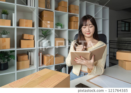 Young Woman Taking Online Sales Orders in Modern Home Office Surrounded by Shipping Boxes and Shelves Young Woman Taking Online Sales Orders in Modern Home Office Surrounded by Shipping Boxes and Shelves 121764385