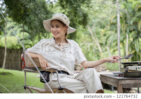 Young Woman Camping and Cooking in the Forest, Enjoying Outdoor Adventure and Nature, Wearing Hat and Casual Clothing 121764732