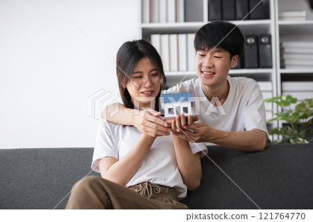 Young Couple Exploring Real Estate Opportunities, Holding a Model House, and Discussing Future Home Purchase Plans in a Modern Living Room Setting 121764770
