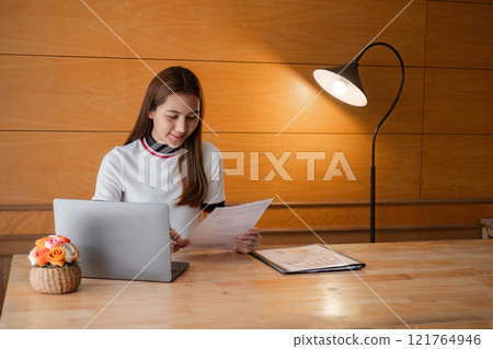 Young Woman Concentrating on Paperwork in a Cozy Room with Modern Decor and Warm Lighting Young Woman Concentrating on Paperwork in a Cozy Room with Modern Decor and Warm Lighting 121764946