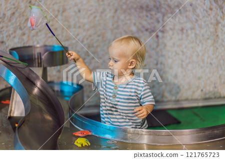 Toddler boy catching toy fish with a net in a vibrant water play area at an entertainment center. A joyful moment of learning, play, and discovery designed to inspire young children 121765723