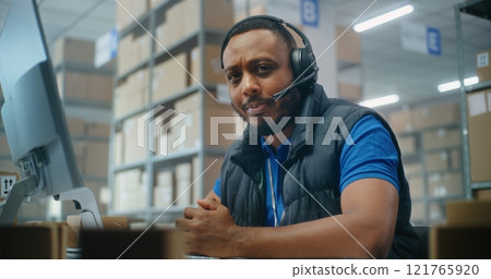 Logistics customer support specialist in headset talks to client, uses computer, looks at camera Logistics customer support specialist in headset talks to client, uses computer, looks at camera 121765920