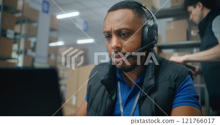 African American retailer or logistics coordinator in headset talks to client, works on laptop in warehouse African American retailer or logistics coordinator in headset talks to client, works on laptop in warehouse 121766017